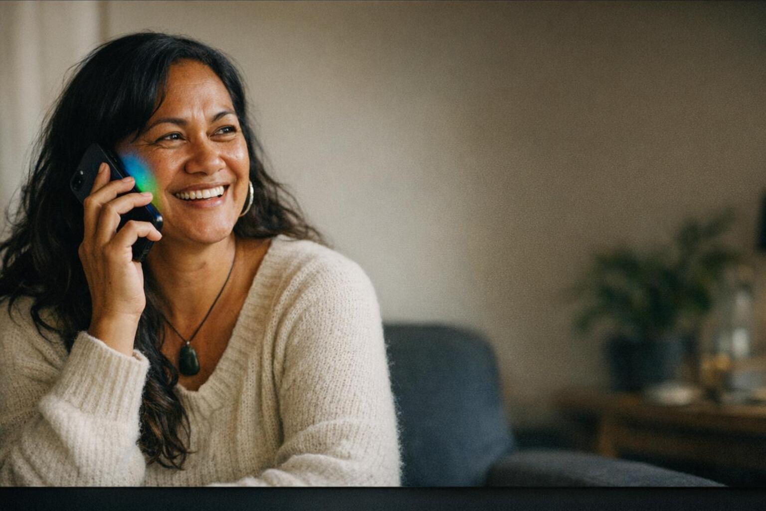 A New Zealand woman on the phone with a warm, genuine smile, mid natural conversation, a soft blue to green glow coming from her phone.